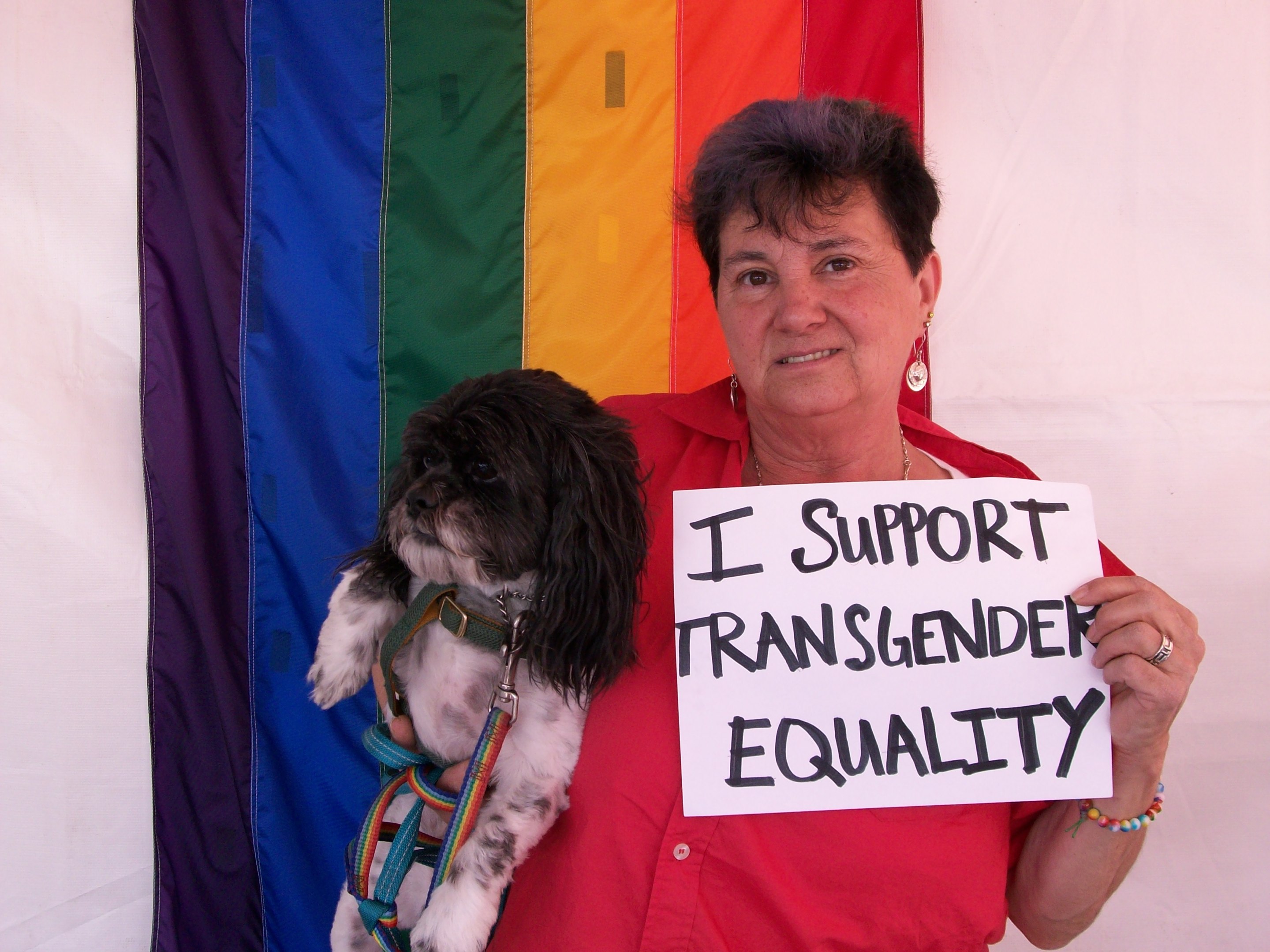 Individual holding dog poses in front of rainbow banner with sign reading "I Support Transgender Equality."