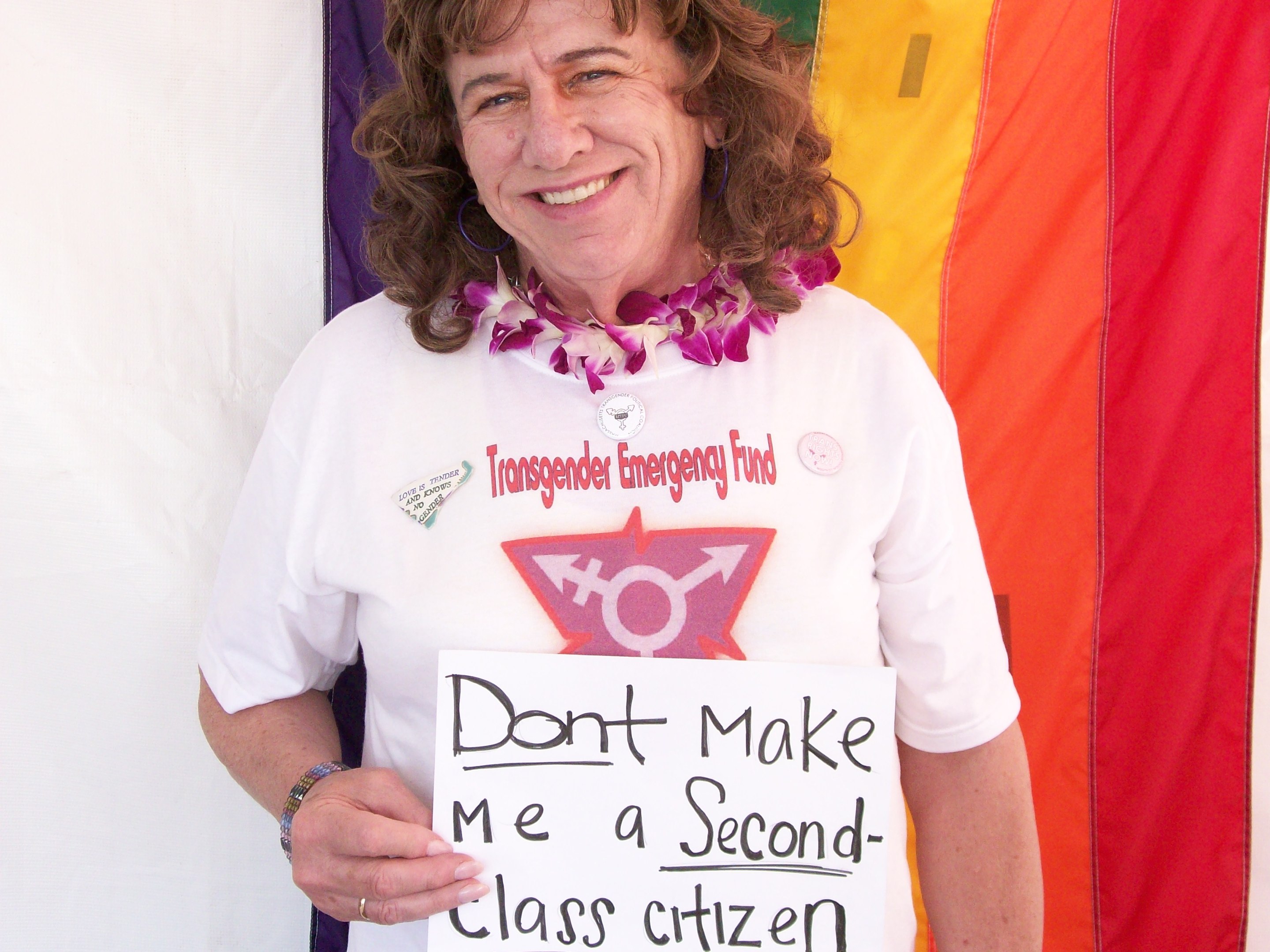 Individual wearing Transgender Emergency Fund shirt smiles in front of rainbow banner with sign reading "Don't Make Me a Second-Class Citizen."
